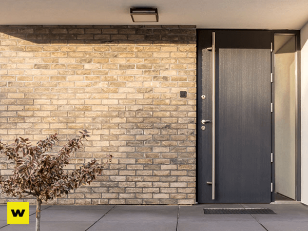 Moderne Haustür aus dunklem Aluminium neben heller Klinkerfassade mit kleiner Pflanze.